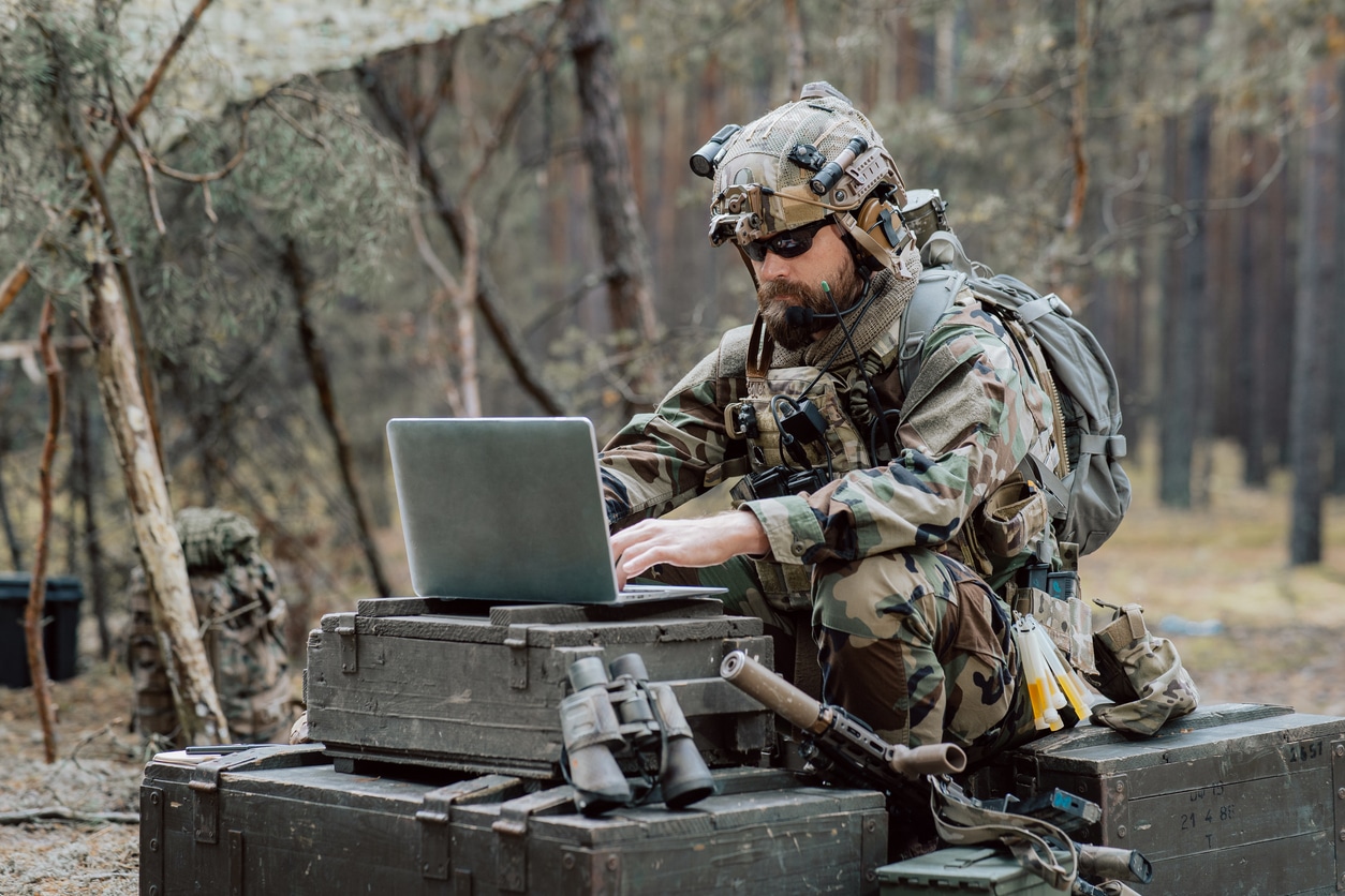 Bearded soldier in uniform sit on military transport crates, analyze data on a laptop and work out tactics at a temporary forest base. In the background, you can see a soldier protecting the base.