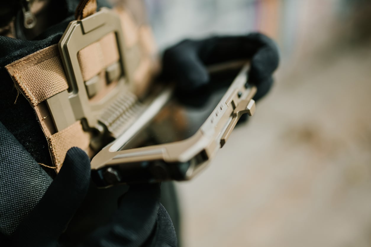 Soldier wearing tactical gloves is using a ruggedized tablet computer attached to their gear, potentially for communication or navigation purposes in a field operation